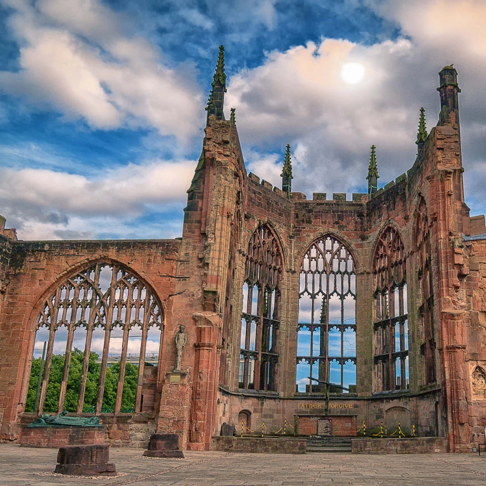 Ruined stone walls and tall Gothic window openings of Coventry Cathedral’s old structure under a partly cloudy sky, showing the shell of the historic cathedral destroyed in World War II with no roof and ornate stone tracery still standing as a memorial.
