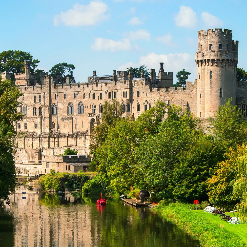 Exterior view of Warwick Castle in Warwickshire, England — a large, medieval stone fortress with tall towers and crenellated walls set on high ground, overlooking surrounding greenery and historic grounds, showing its classic battlements and Norman-era architecture.