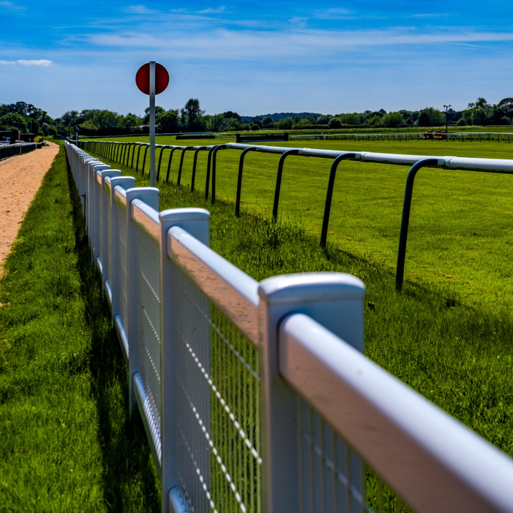 A wide view of Warwick Racecourse in Warwickshire, England, showing the grassy horse racing track curving around with fences or jumps, grandstands and spectator areas along the side, and open countryside beyond, capturing the racecourse layout and event setting typical of National Hunt horse racing.